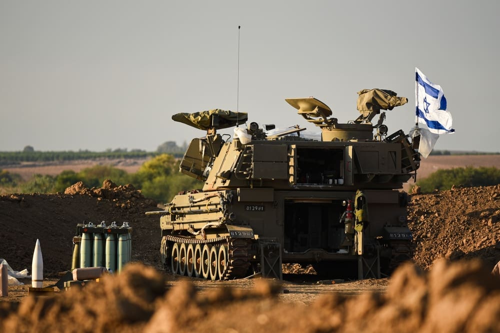 An IDF M109 artillery cannon is positioned on a dirt mound with an Israeli flag, prepared to fire