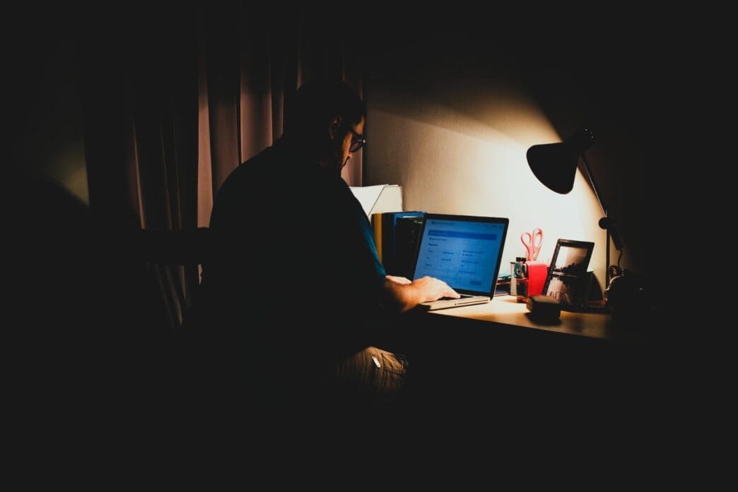 A man sits in a dark room, illuminated by a desk lamp as he uses his laptop to navigate illegal content