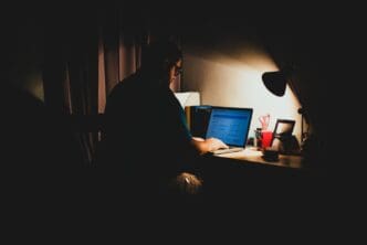 A man sits in a dark room, illuminated by a desk lamp as he uses his laptop to navigate illegal content
