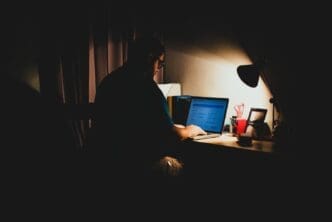 A man sits in a dark room, illuminated by a desk lamp as he uses his laptop to navigate illegal content