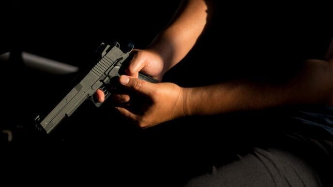 A person is sitting in a dark room holding a handgun, with a focus on their hand