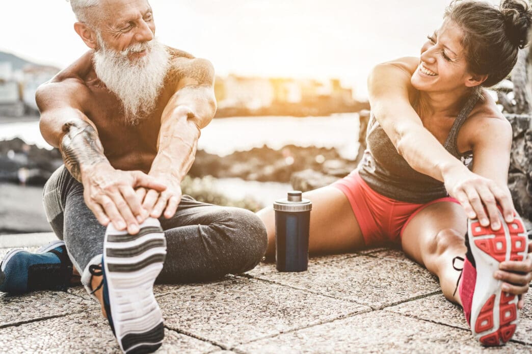 A smiling older man with a white beard and a young woman stretch together on a paved surface near the water