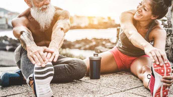 A smiling older man with a white beard and a young woman stretch together on a paved surface near the water