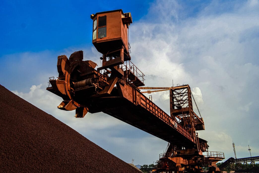 A large, rusty iron ore stacker-reclaimer machine against a cloudy sky