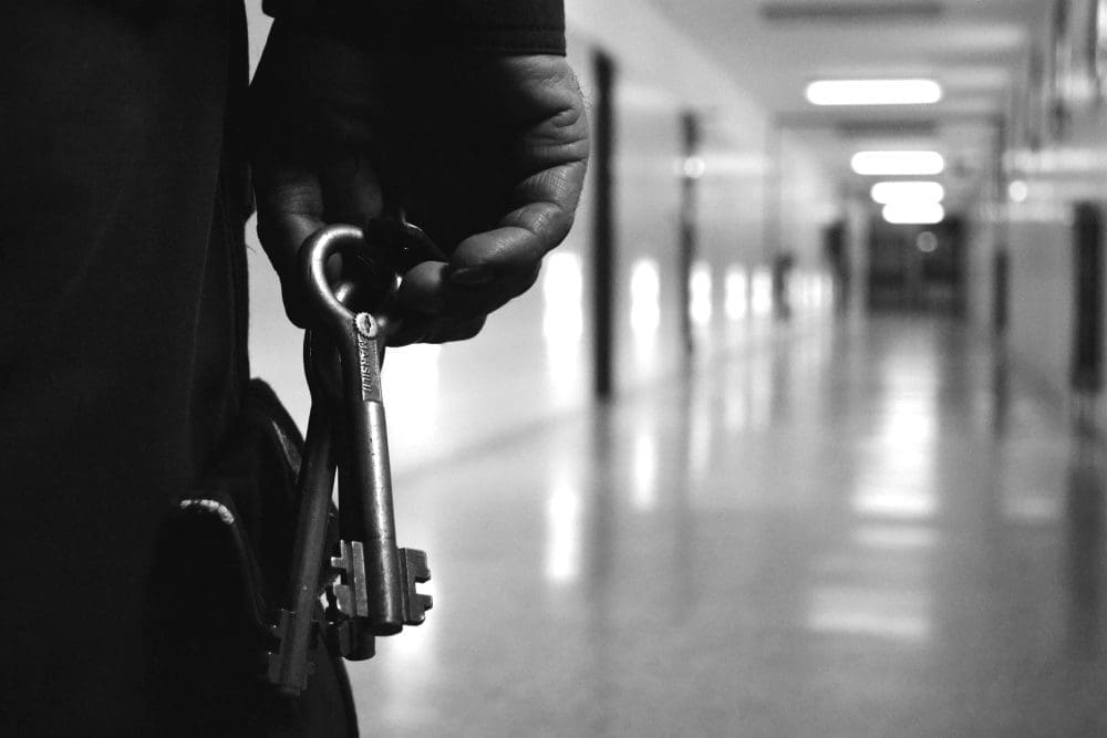 A jailer's hand holding a large ring of keys with a prison hallway in the background