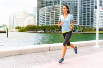 A smiling woman jogs along a waterfront path in downtown Miami with modern buildings behind her