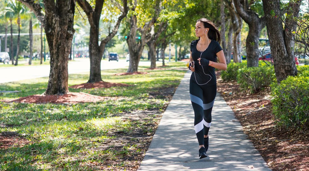 A woman with a ponytail and headphones jogs on a sidewalk in a leafy park