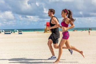 A man and a woman jogging barefoot on a sandy beach next to the ocean on a sunny day
