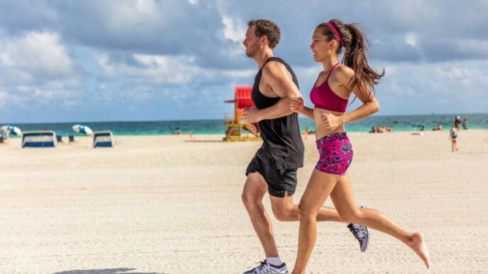 A man and a woman jogging barefoot on a sandy beach next to the ocean on a sunny day
