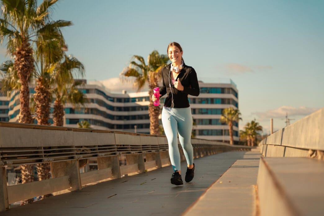 A woman jogs on a bridge with headphones and a water bottle