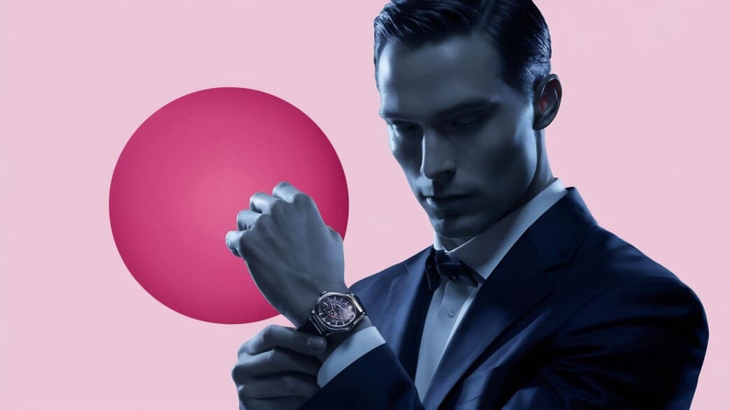 A man in a suit adjusts his watch against a pink and red backdrop