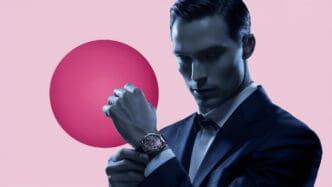 A man in a suit adjusts his watch against a pink and red backdrop