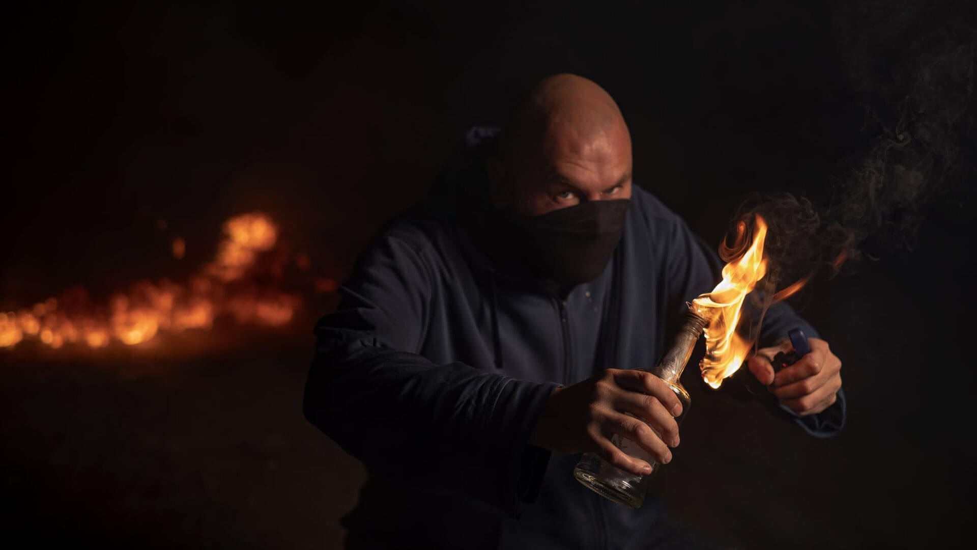 Man in mask and hoodie lighting a Molotov cocktail in dark surroundings