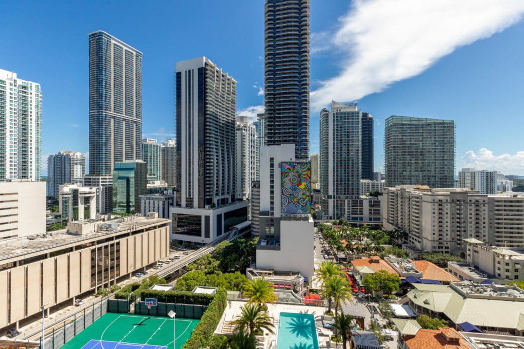 A high-angle view of the Miami skyline, showing tall buildings, a monorail track, a basketball court, and a swimming pool in the foreground