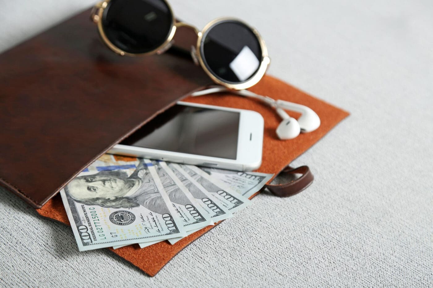 A brown leather folio holding a smartphone, earbuds, and US dollar bills