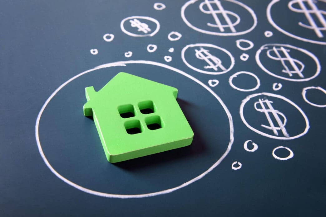 Green house model surrounded by dollar signs drawn on a dark surface