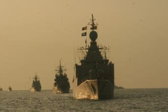 A line of military ships sailing on a choppy sea at dusk, with the largest warship in the foreground