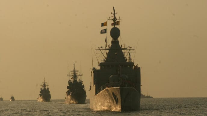 A line of military ships sailing on a choppy sea at dusk, with the largest warship in the foreground
