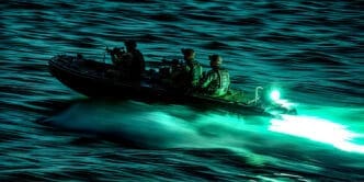 Three armed soldiers in a military-style rigid inflatable boat speed across dark, choppy water at night