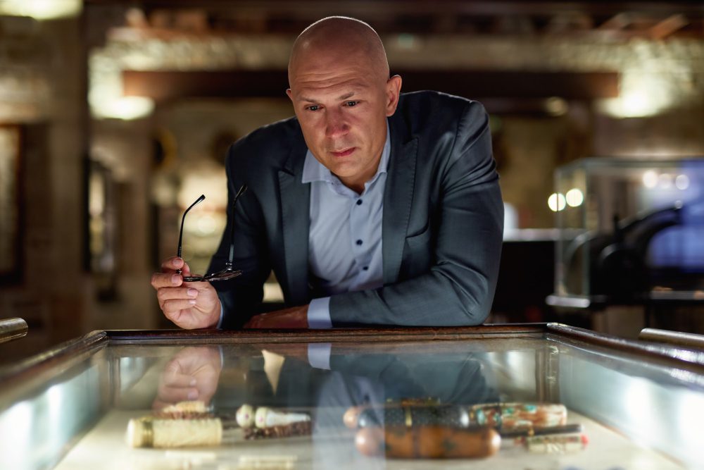 A bald man in a suit holds a pair of glasses and looks intently into a museum display case