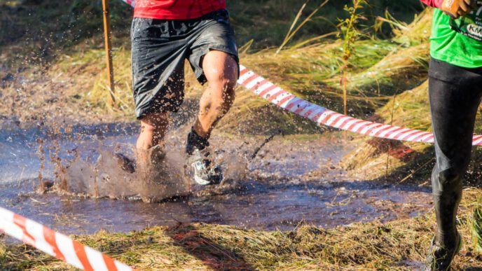 The legs of two competitors running and splashing through a muddy water obstacle during a race