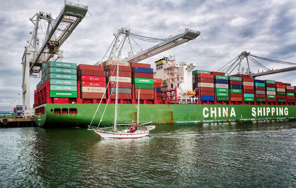 A small sailboat passes in front of a giant cargo ship and cranes at the Port of Oakland