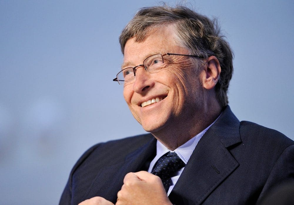 Bill Gates, a man with glasses and graying hair, smiles while wearing a suit