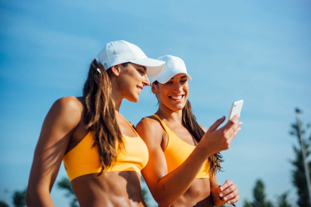 Two fit women in yellow sports bras and white hats smile as they look at a smartphone