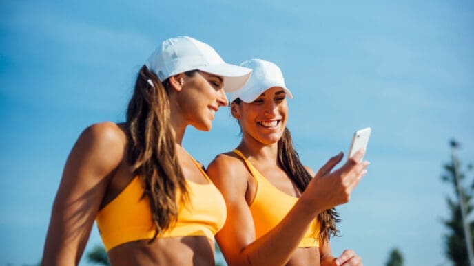Two fit women in yellow sports bras and white hats smile as they look at a smartphone