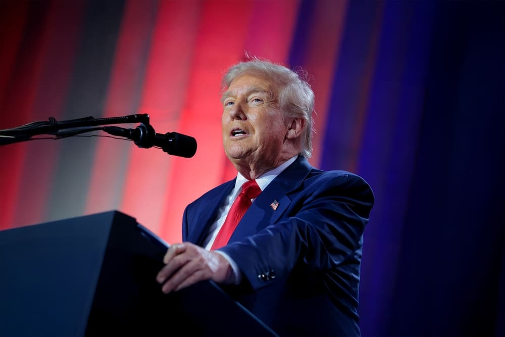 President Donald Trump stands at a podium with a microphone, delivering remarks at a summit with red and blue lighting in the background