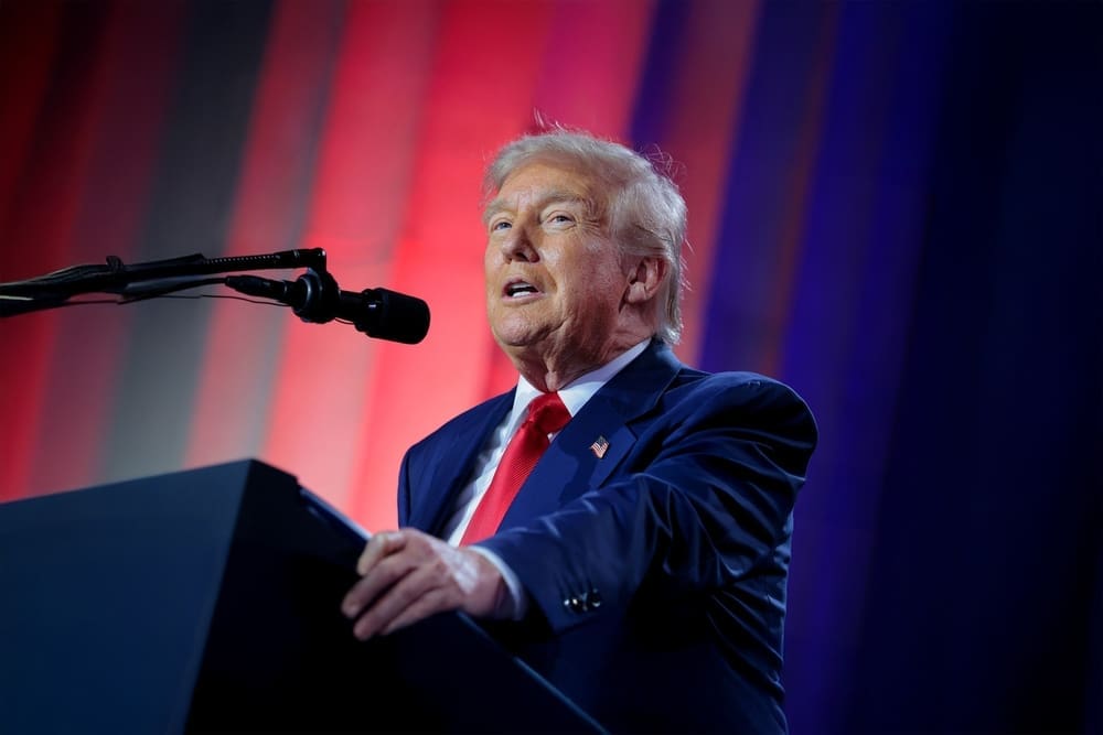 President Donald Trump stands at a podium with a microphone, delivering remarks at a summit with red and blue lighting in the background