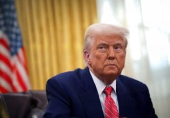 A portrait of President Donald Trump seated in the Oval Office, with a serious expression, and an American flag behind him