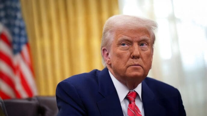 A portrait of President Donald Trump seated in the Oval Office, with a serious expression, and an American flag behind him