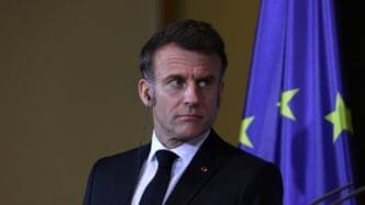 Emmanuel Macron, a man in a suit, stands with a serious expression in front of a European Union flag