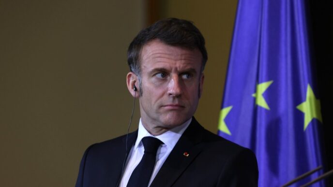Emmanuel Macron, a man in a suit, stands with a serious expression in front of a European Union flag