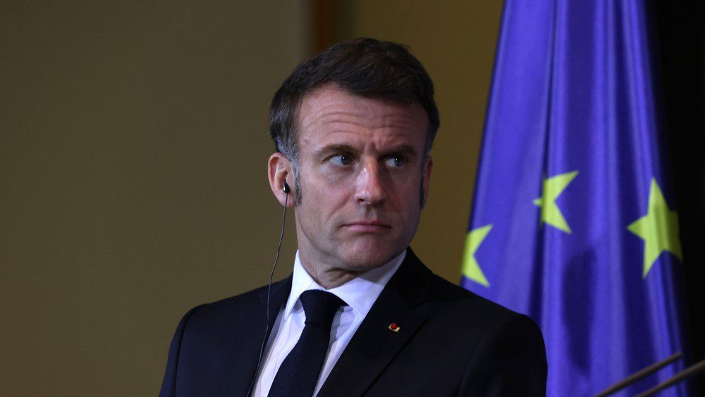 Emmanuel Macron, a man in a suit, stands with a serious expression in front of a European Union flag