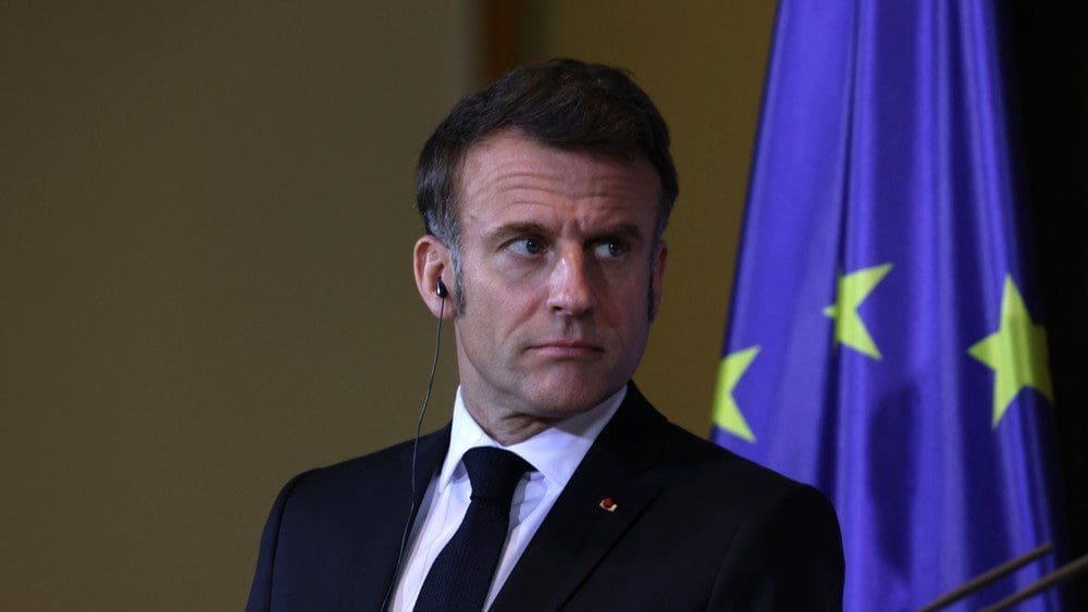 Emmanuel Macron, a man in a suit, stands with a serious expression in front of a European Union flag