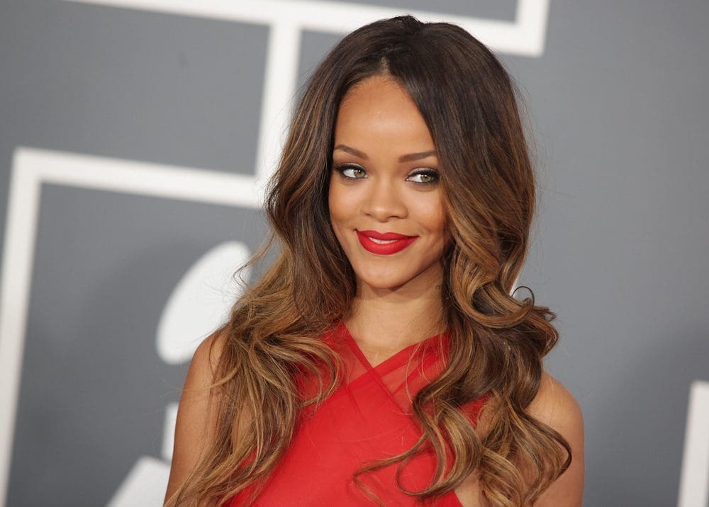 Rihanna with long, wavy brown hair, wearing a red dress, smiles at the camera