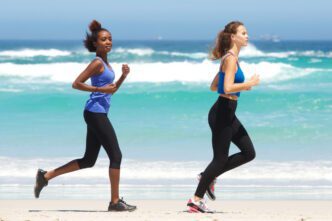 Two women in athletic wear running side-by-side on a sunny beach near the ocean