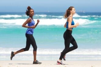 Two women in athletic wear running side-by-side on a sunny beach near the ocean