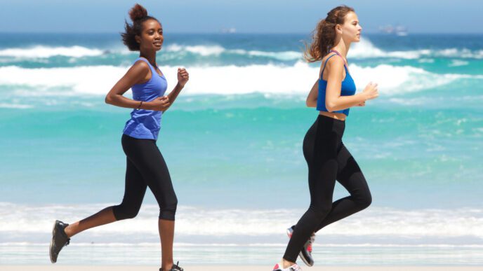 Two women in athletic wear running side-by-side on a sunny beach near the ocean