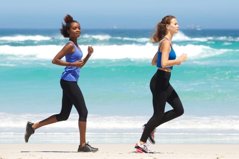 Two women in athletic wear running side-by-side on a sunny beach near the ocean