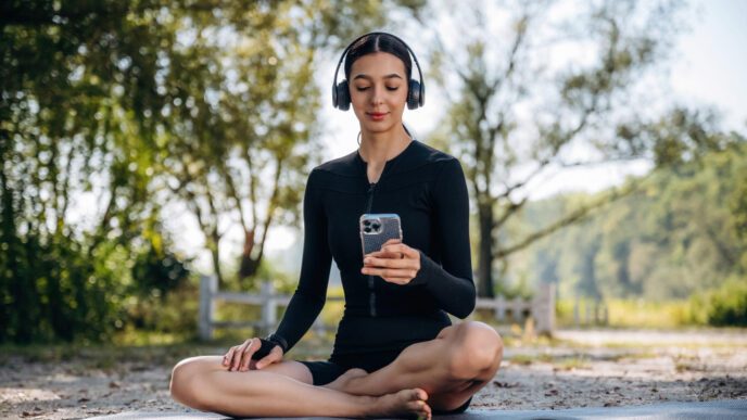 A woman in athletic wear is meditating outdoors with headphones and a smartphone
