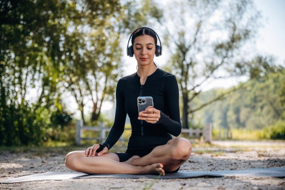 A woman in athletic wear is meditating outdoors with headphones and a smartphone