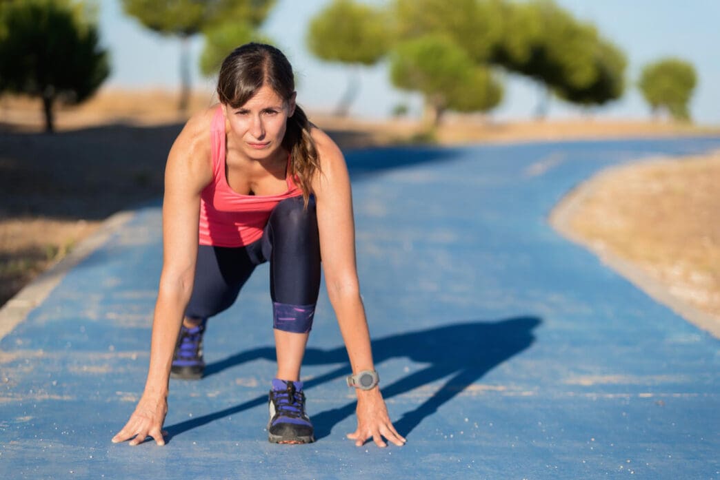 A woman in athletic wear is in a low running start on a blue path with a determined expression