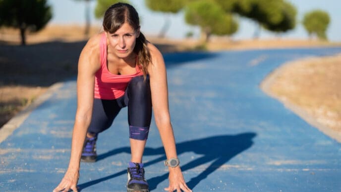 A woman in athletic wear is in a low running start on a blue path with a determined expression