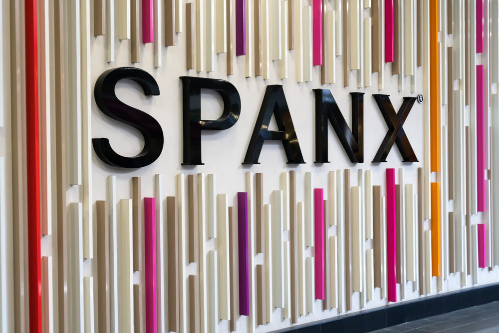 The black SPANX logo is mounted on a textured wall with colorful vertical stripes