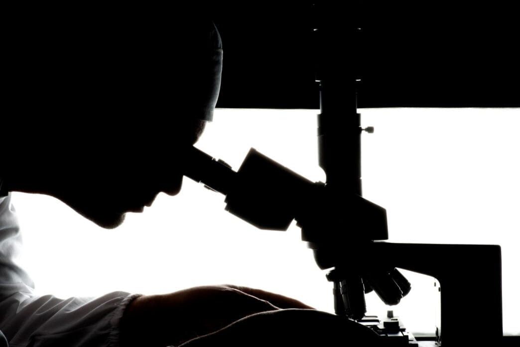 The silhouette of a person in a lab coat looks through a microscope against a bright white background