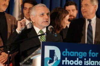 Senator Jack Reed speaks at a podium with a microphone to a crowd in front of a blue backdrop