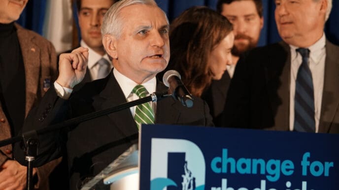 Senator Jack Reed speaks at a podium with a microphone to a crowd in front of a blue backdrop